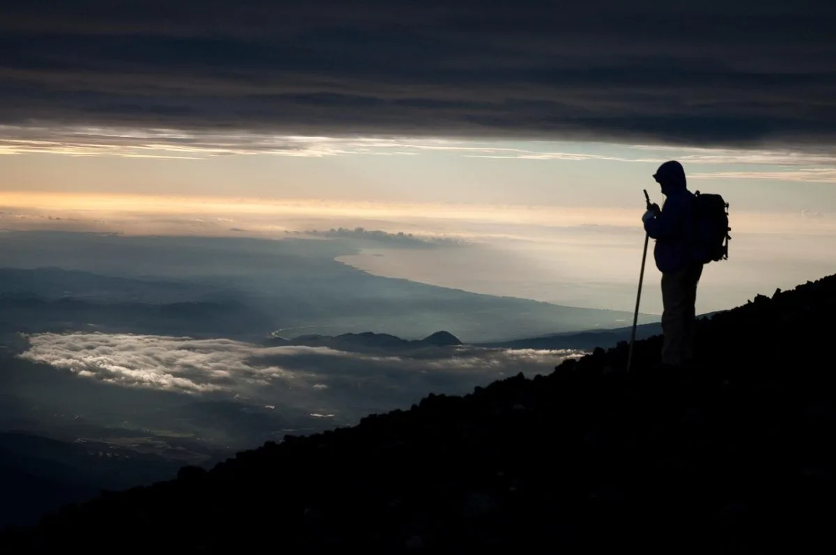 Silhouette of a hiker ascending Mount Fuji at dawn, overlooking a sea of clouds and the distant coastline under dramatic skies.