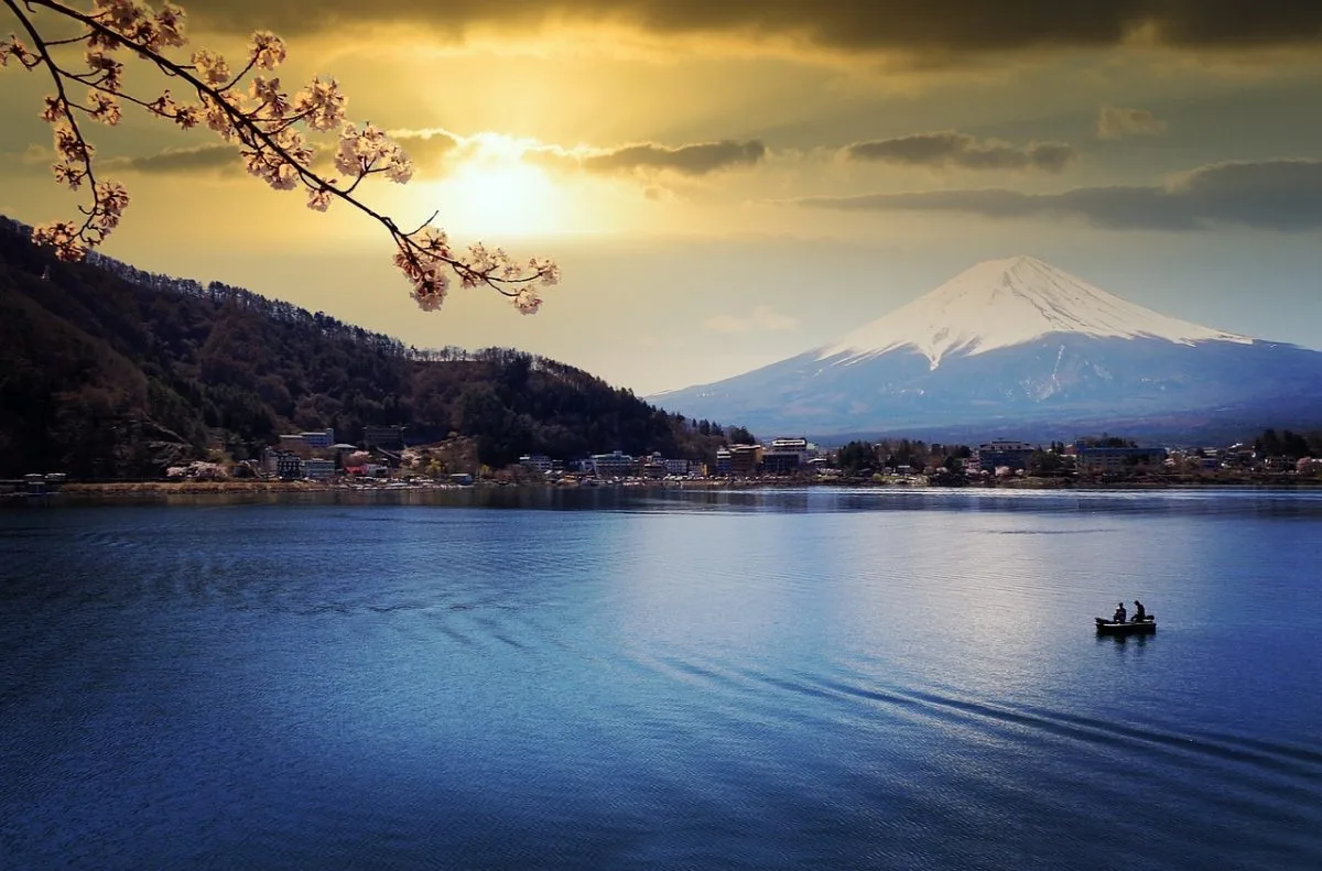 Scenic view of Mount Fuji with snow-capped peak at sunset, cherry blossoms in the foreground, and a boat on Lake Kawaguchiko – a peaceful moment capturing the natural beauty of Japan's iconic volcano.