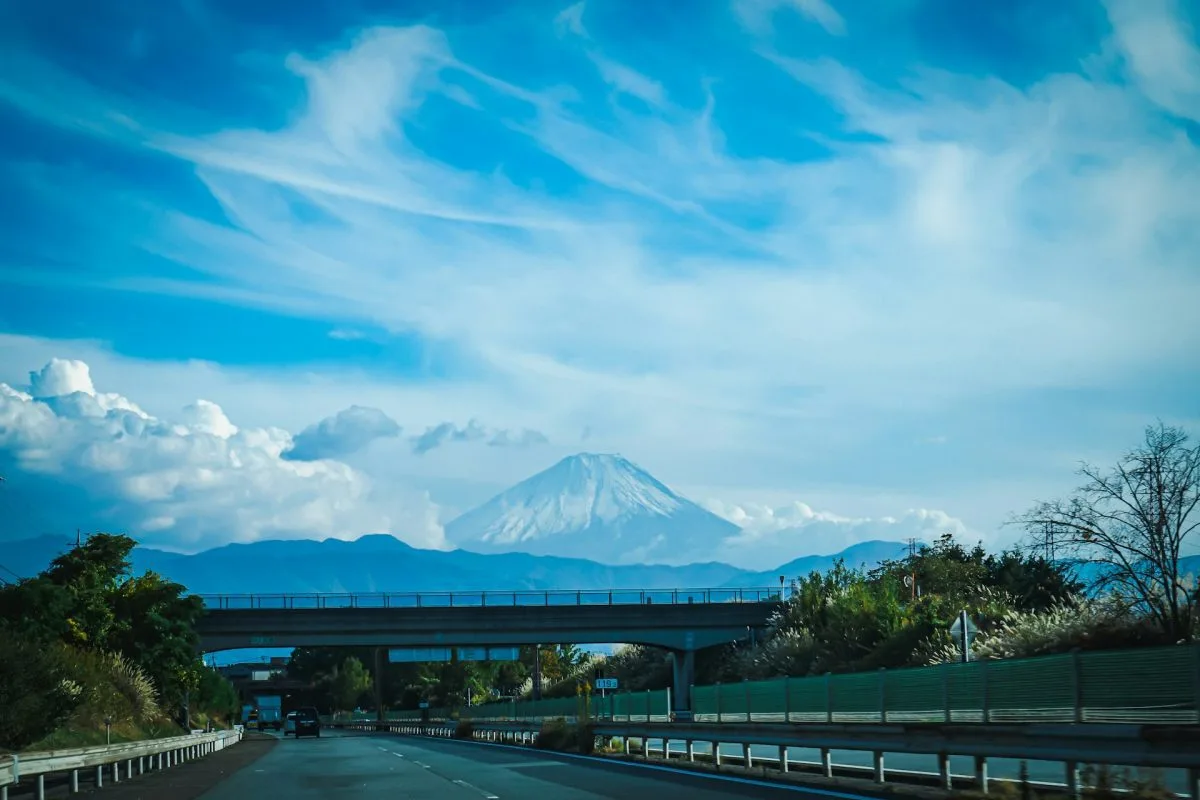 Scenic view of Mount Fuji from a car on a highway, showcasing a travel perspective with roads, an overpass, and the iconic mountain against a blue sky.