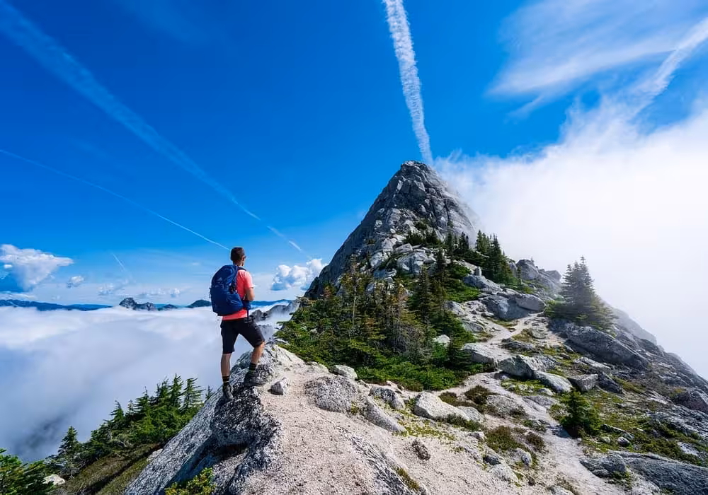 Daniel Reed standing on a rocky mountain ridge, looking out at a majestic peak surrounded by a sea of clouds under a vibrant blue sky, embodying authentic adventure travel.
