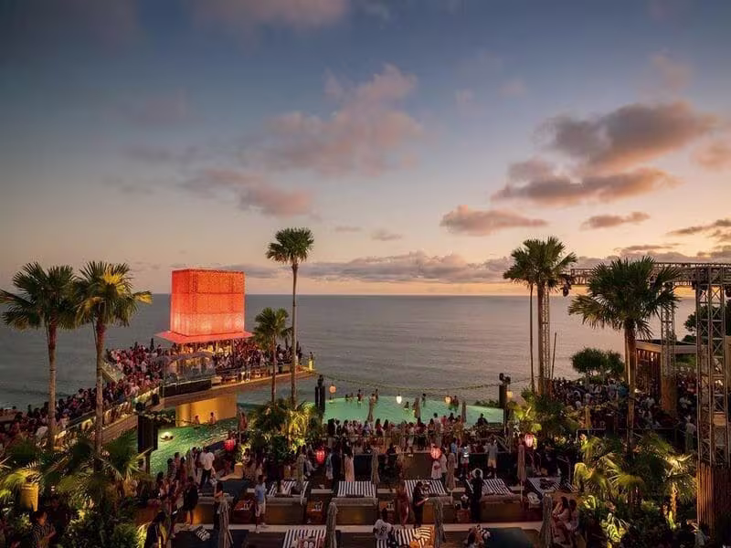 Sunset party atmosphere at Savaya Bali in Uluwatu, showcasing guests around the glowing Cube bar, main pool, and various lounges overlooking the sea as evening approaches.