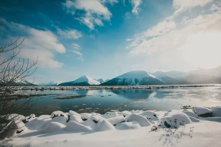 Sunlight on a tranquil Alaskan lake with snow-covered banks and distant mountains, highlighting the peaceful beauty when considering the best time to visit Alaska.