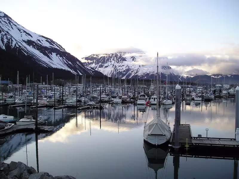 Fishing boats moored in a tranquil Alaskan port at the base of snowy mountains, representing the start of a fishing trip.