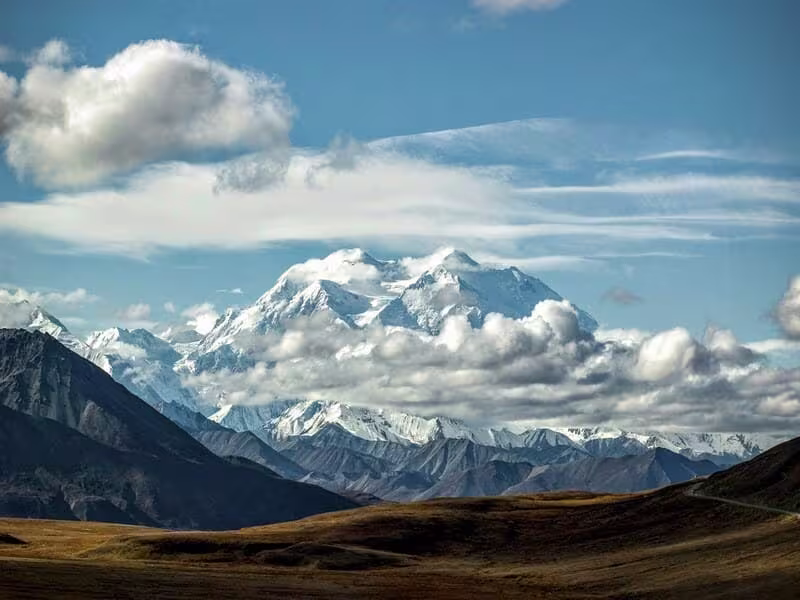 Denali (Mount McKinley) towering over the Alaskan tundra, with its snow-covered peaks partially obscured by clouds under a blue sky.
