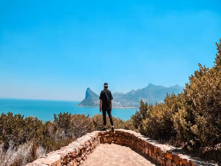 Daniel Reed, wearing a hat, stands on a stone viewpoint overlooking a stunning coastal scene with turquoise water and distant mountains, representing a moment of travel discovery.