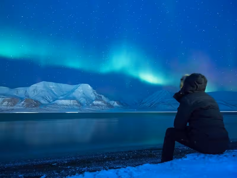 Person watching the vibrant green and teal Northern Lights (aurora borealis) over a snowy Alaskan landscape with mountains and reflective water.