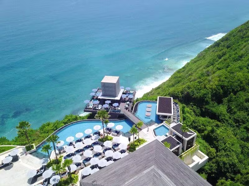High-angle daytime view of Savaya Bali, showing its multi-tiered infinity pools, numerous sun loungers with umbrellas, the iconic Cube bar, and the expansive turquoise Indian Ocean meeting a lush green cliffside.