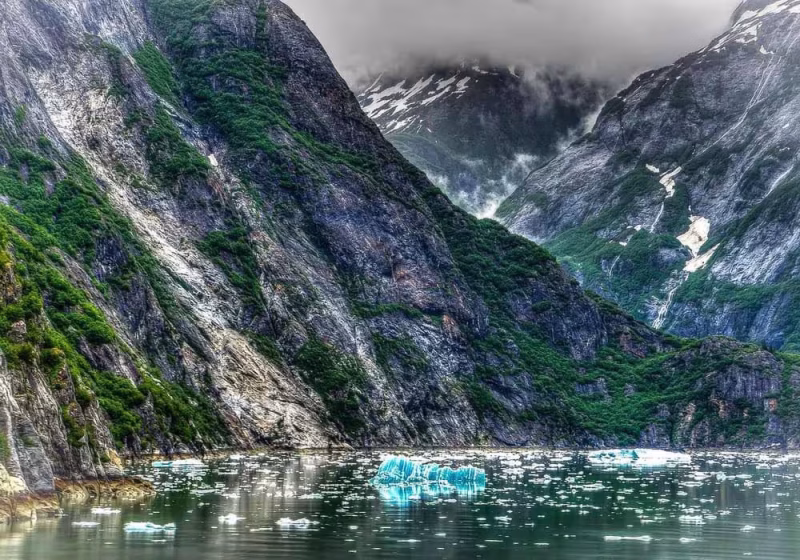 Scenic view of Alaska in spring, featuring floating ice in a fjord, misty mountains with snow, and early green vegetation on the slopes.