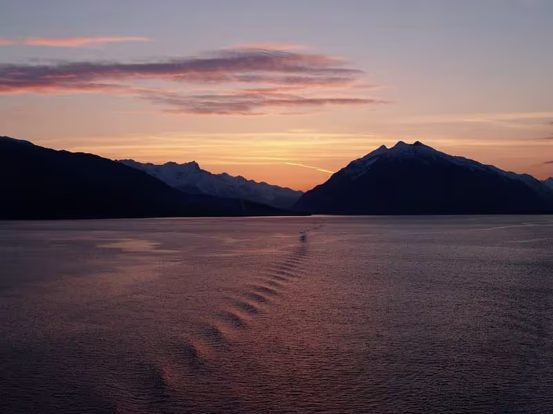 Sunset over a wide Alaskan bay or fjord, with mountains in silhouette and colorful sky reflected on the water – an ideal time to visit Alaska.