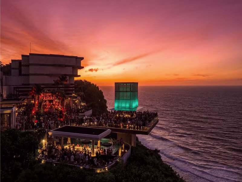 Vibrant sunset illuminates Savaya Bali, with crowds gathered on the multi-level cliffside decks and the iconic Cube glowing turquoise against the twilight sky over the Indian Ocean.